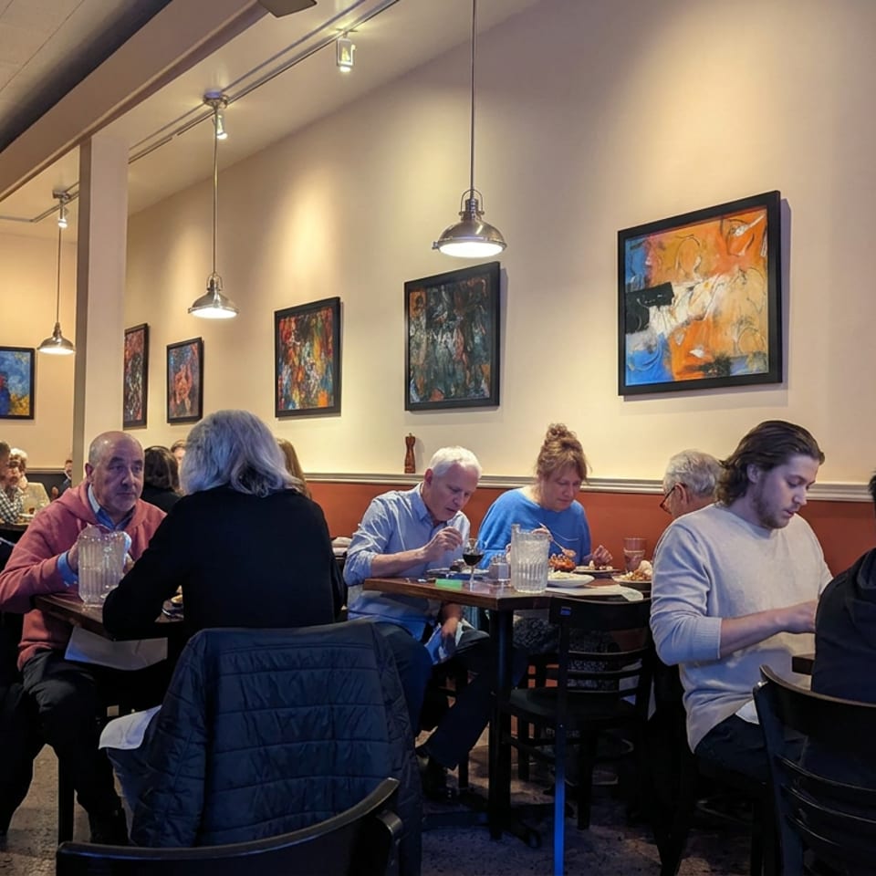 Busy restaurant dining room with patrons seated at tables beneath pendant lights and colorful abstract artwork.
