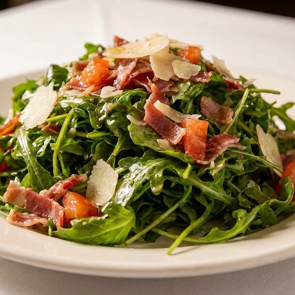 Arugula salad with crispy bacon, tomatoes, and shaved Parmesan served on a white plate.