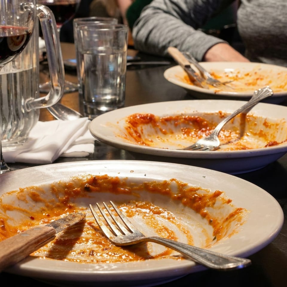 Three nearly empty plates with tomato sauce, utensils, water glasses, grated cheese, and red wine on a restaurant table.