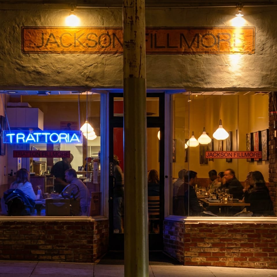Nighttime street view of Jackson Fillmore Trattoria, with diners visible through warmly lit windows and a blue neon sign.