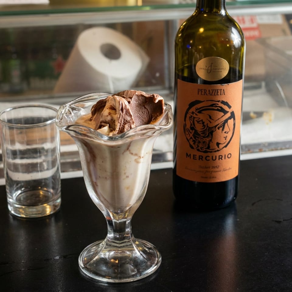 Chocolate gelato in a glass dessert dish sits beside an empty tumbler and Perazzeta Mercurio wine bottle on a restaurant counter.