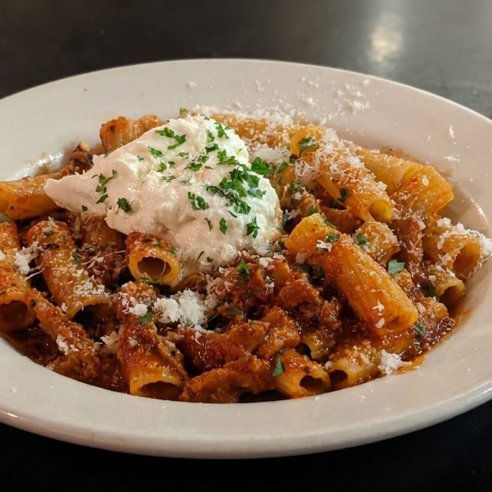 Rigatoni pasta in tomato meat sauce topped with creamy cheese, grated Parmesan, and parsley on a white plate.