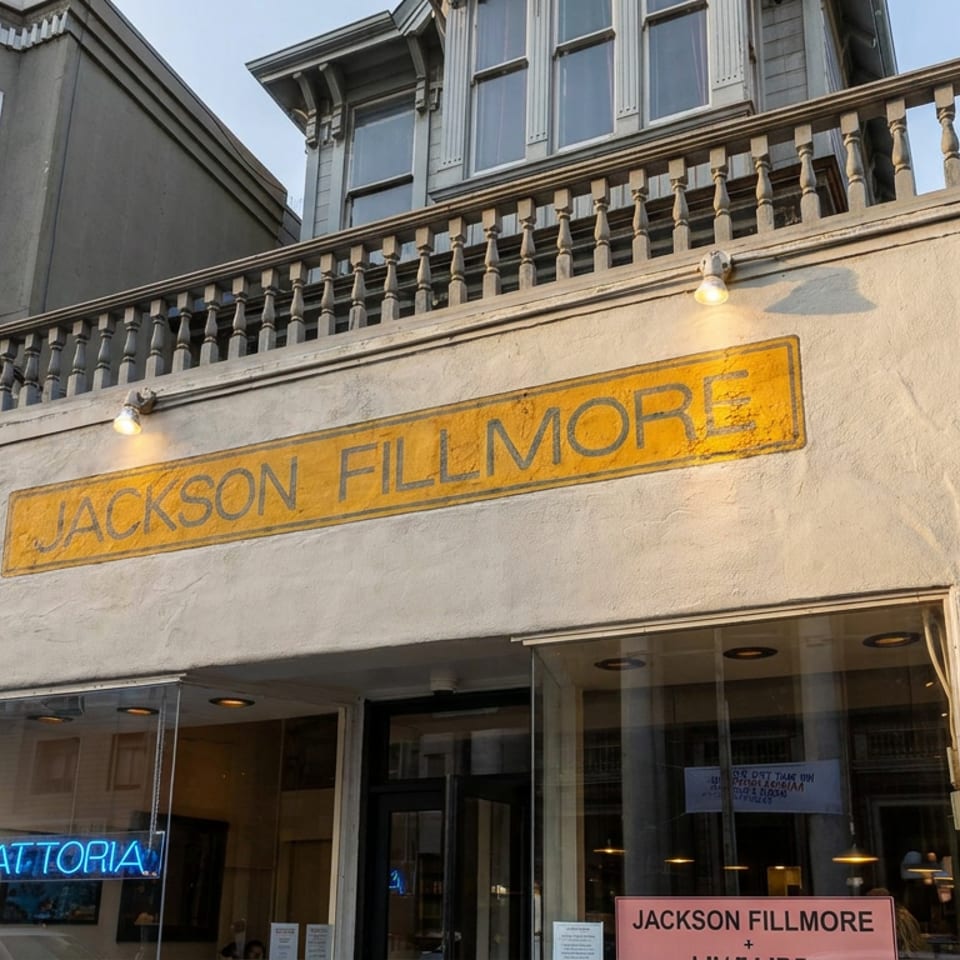 Street-level view of Jackson Fillmore trattoria storefront with yellow signage, neon window lettering, and residential architecture above.